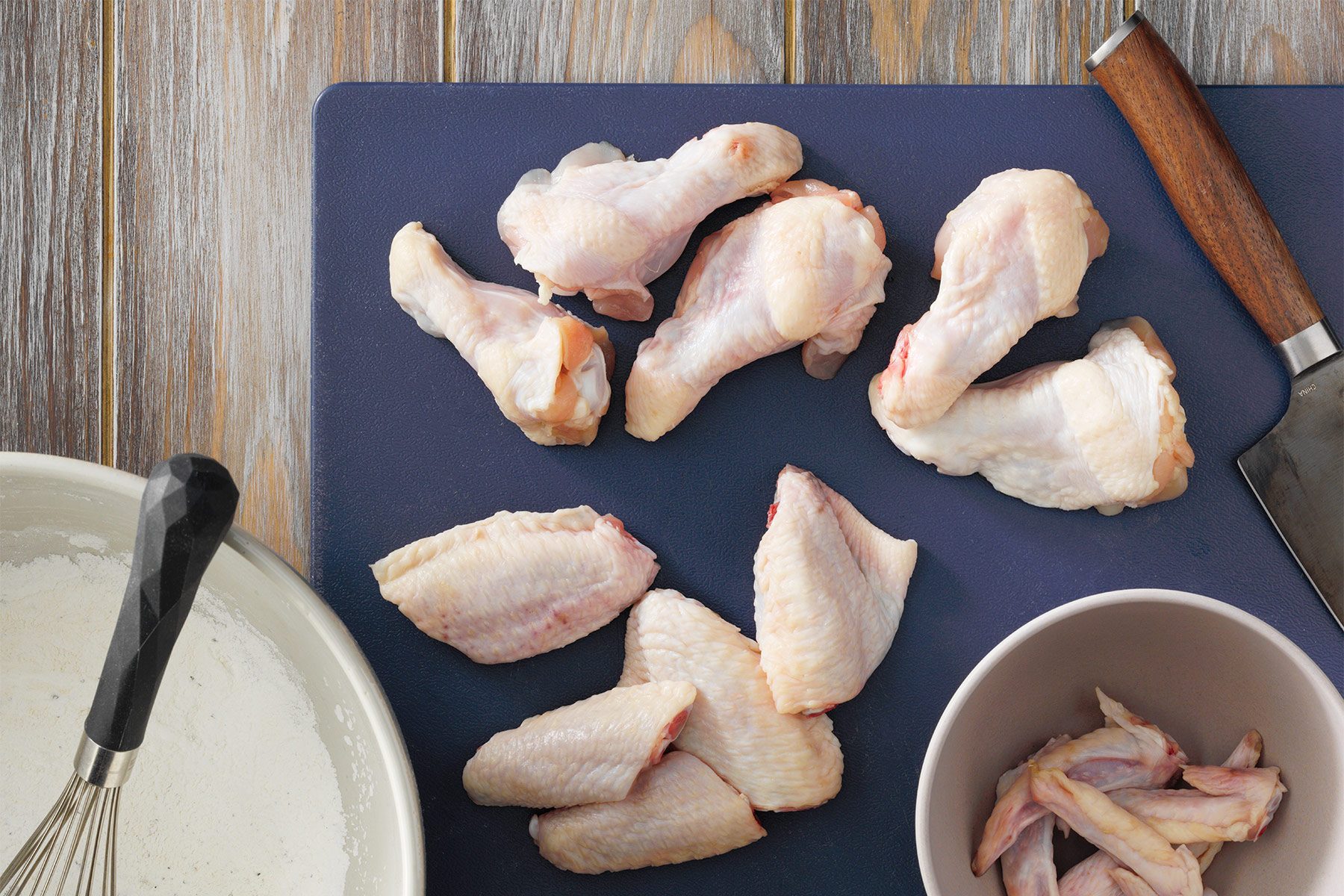 Raw chicken drumsticks and wings are shown on a dark cutting board placed on a wooden surface. Some pieces are also in a bowl and a few are next to a white pan. The ingredients are ready for preparation.