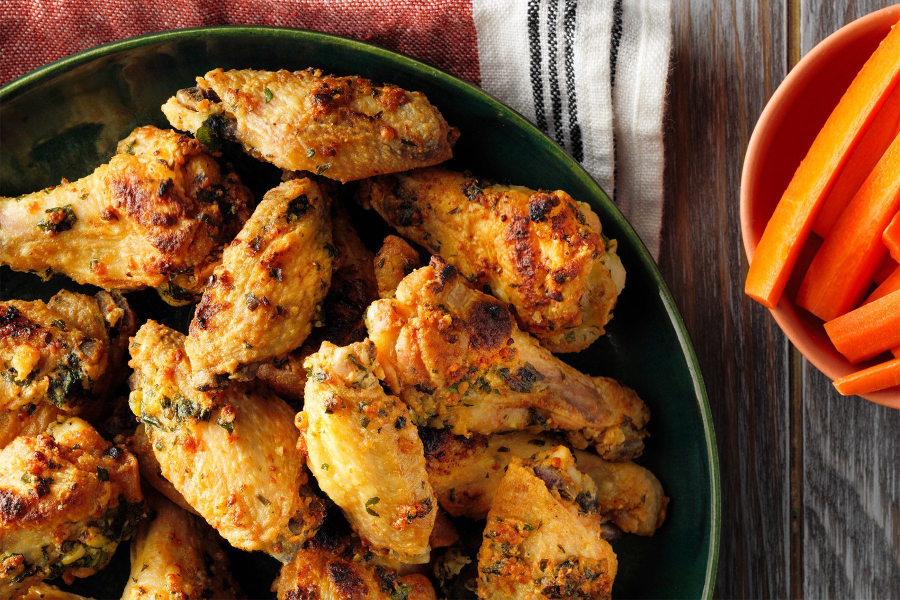 A close-up of a green bowl filled with crispy, golden-brown baked chicken wings, garnished with herbs. The bowl is placed on a cloth napkin with stripes in the background. The chicken wings have a textured, flavorful-looking coating.
