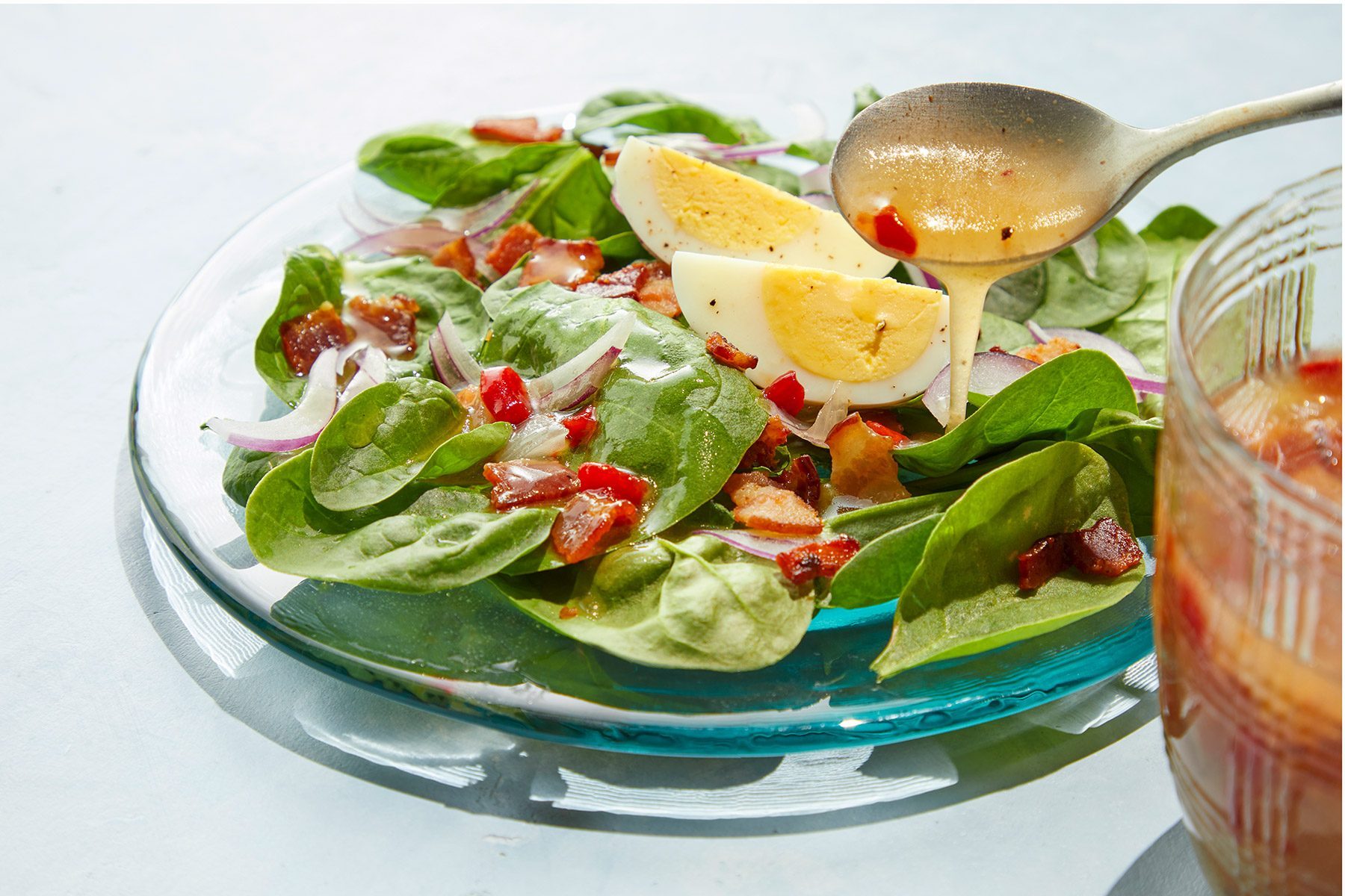 3/4 angle view shot of Hot Bacon Dressing; in a cup; pour over salad; plate; spoon; light blue background