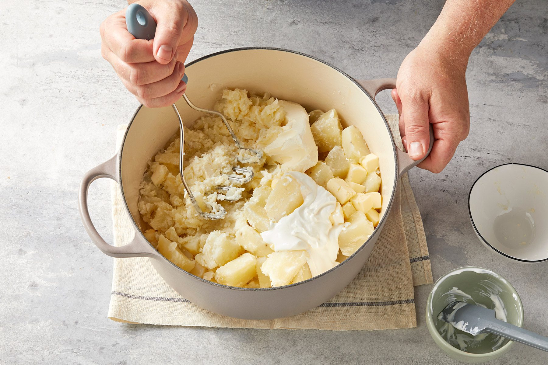 A person is using a potato masher to mash cooked potatoes in a large pot. The pot is placed on a countertop with a small bowl and spoon nearby. The mashed potatoes are mixed with white ingredients, possibly butter and cream.