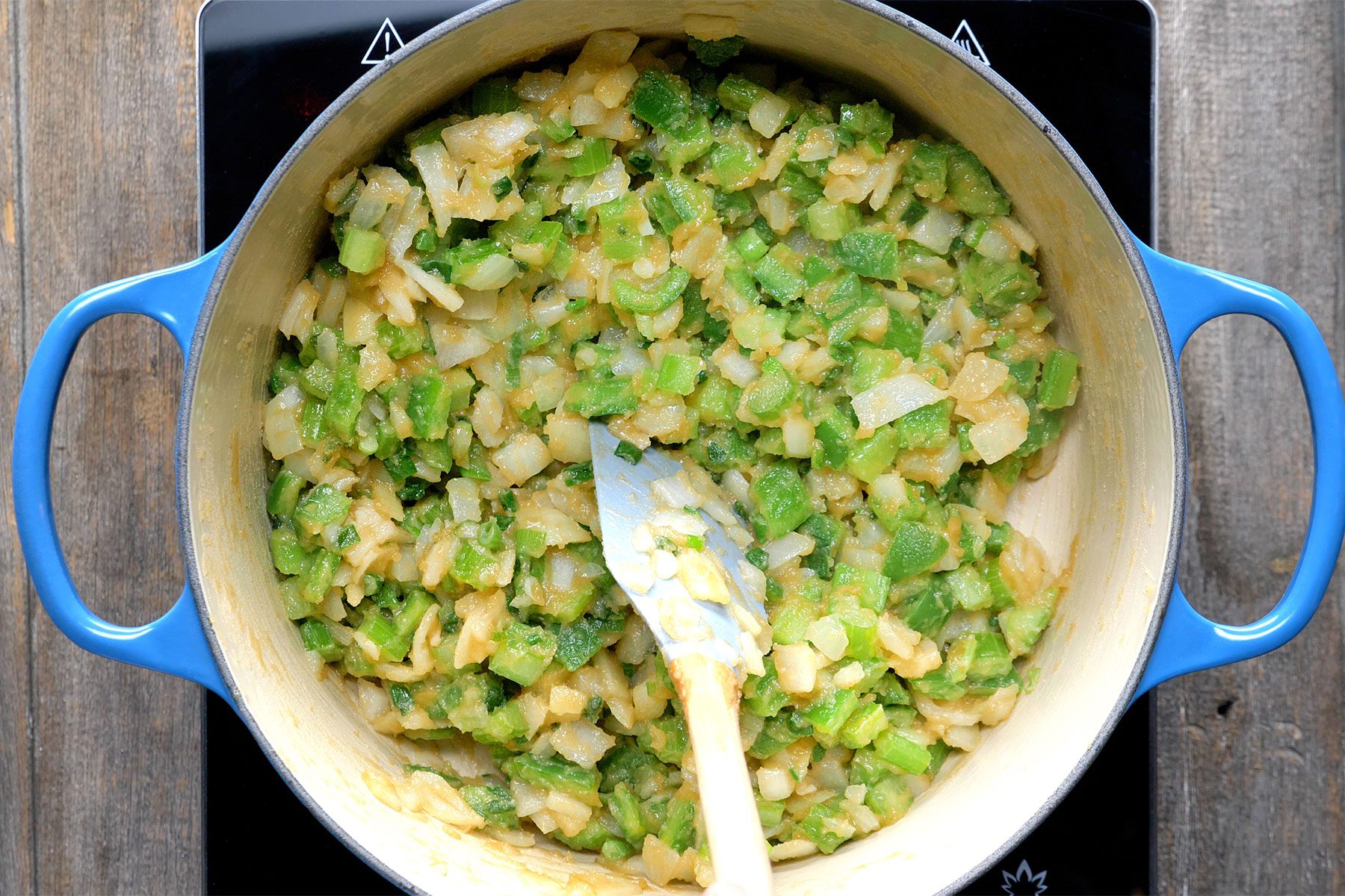 Overhead shot of added the onion; celery; green pepper and green onions in a dutch oven; cook and stir for 5 minutes; spatula; wooden background;