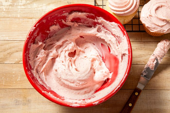 overhead shot of strawberry frosting in large red bowl and knife placed on right side and cup cakes on wired rack on wooden background