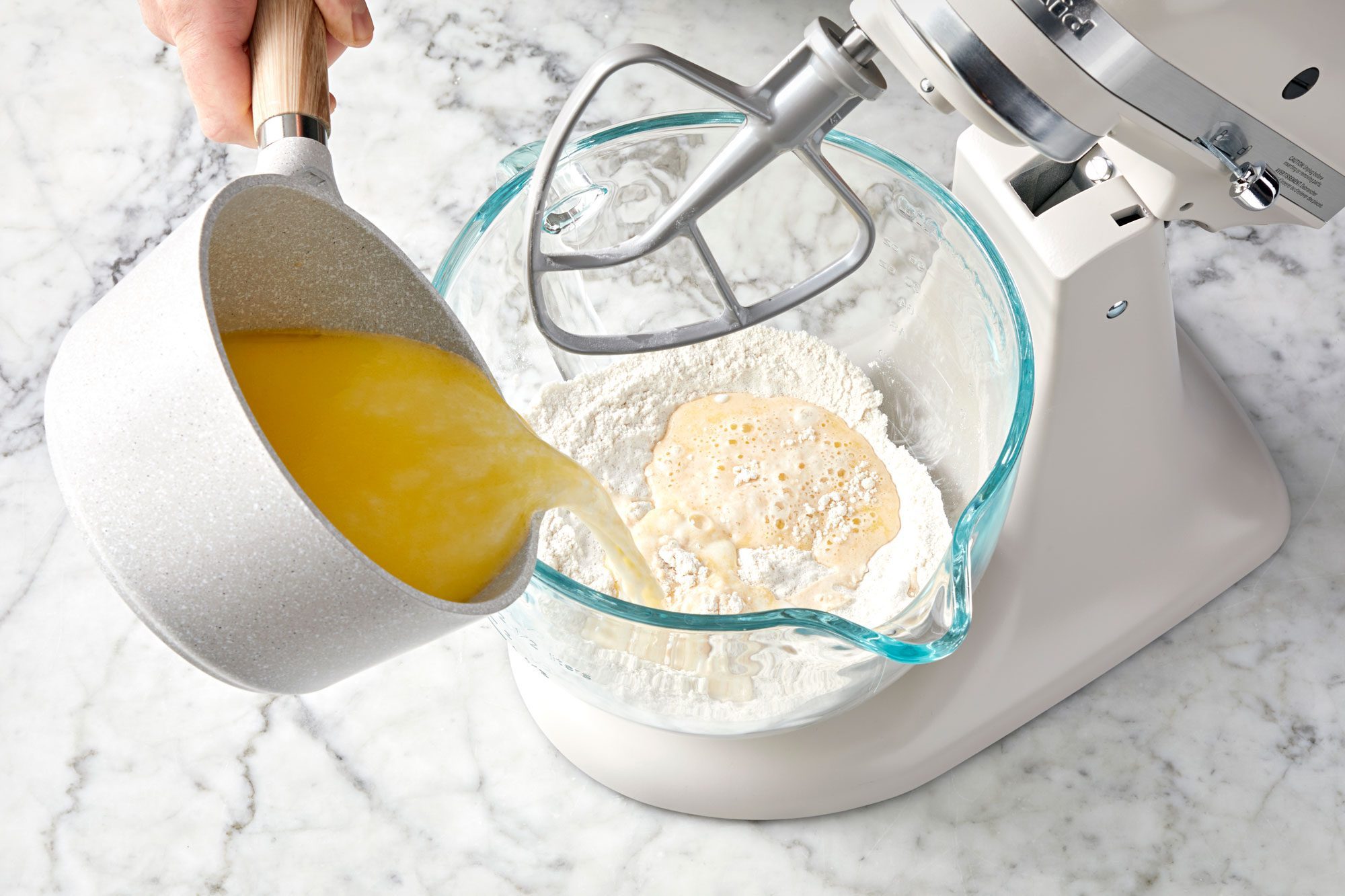 wide shot of pouring yellow mixture into a glass bowl; stand mixer; marble surface;