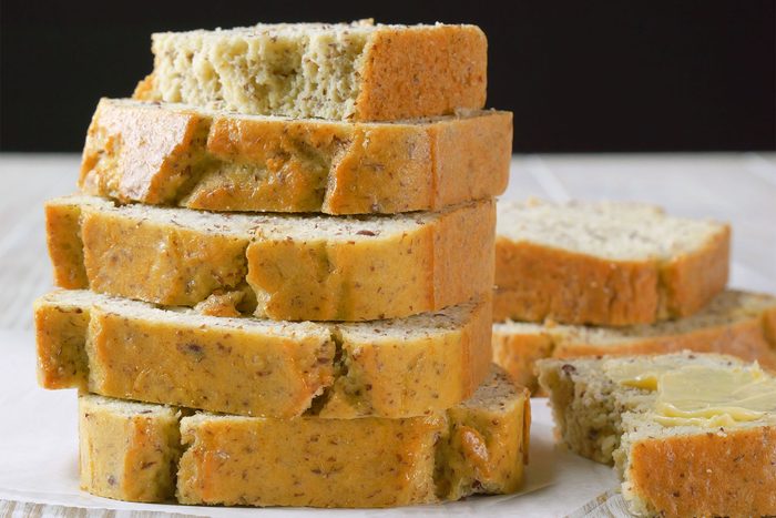 A neatly stacked pile of several slices of moist, homemade banana bread with a golden-brown crust and speckled interior, suggesting the presence of nuts or seeds, on a light-colored plate. A partially buttered slice rests beside the stack.