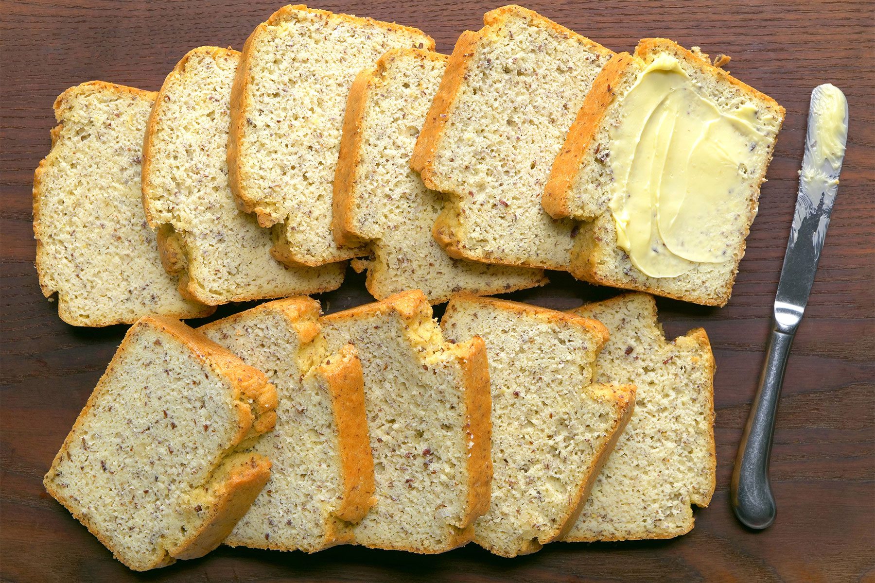Slices of banana bread arranged on a wooden surface. One slice is spread with butter, and a butter knife with some butter on it is placed next to it. The bread has a slightly golden crust and a speckled interior, indicating the presence of bananas and possibly nuts.