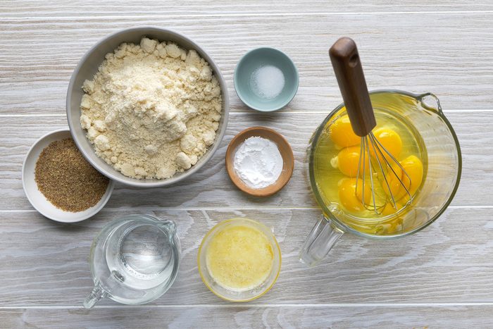 Top-down view of baking ingredients on a wooden surface, including a bowl of flour, a small bowl of salt, a bowl of eggs being whisked, a bowl of baking powder, ground flax seeds, a glass jug of water, and a container of melted butter.