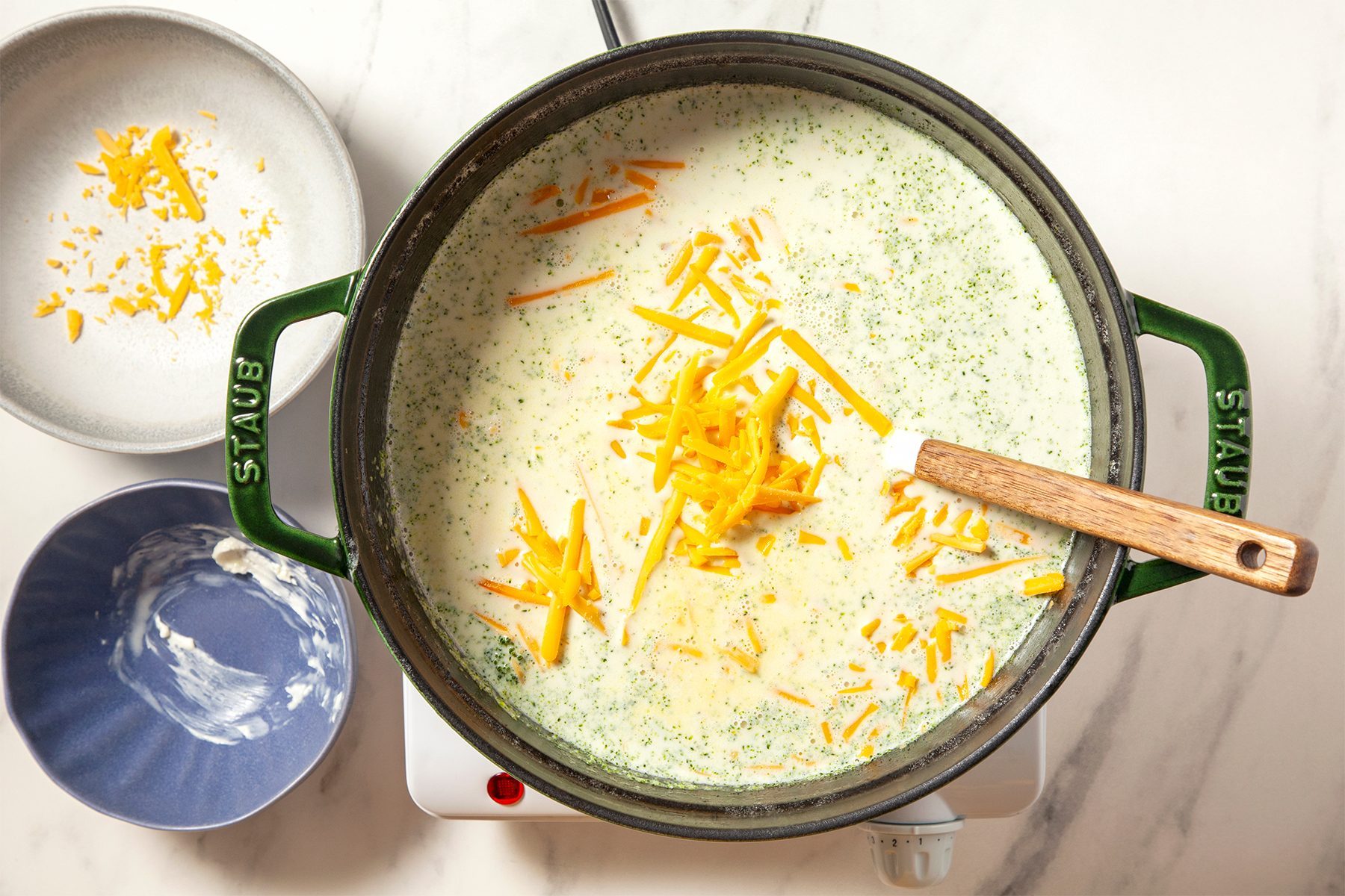 An overhead shot of a pot on a stovetop containing creamy broccoli cheese soup with shredded cheddar cheese on top and a wooden spoon resting inside. To the side are two bowls, one with remnants of sauce and the other with more shredded cheddar cheese.