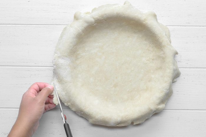 A person's hand is seen trimming the excess dough from the edge of an unbaked pie crust in a pie dish using a pair of scissors.