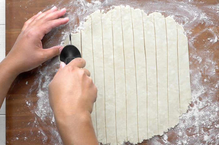 Two hands are cutting dough into strips on a floured wooden surface using a pastry wheel.