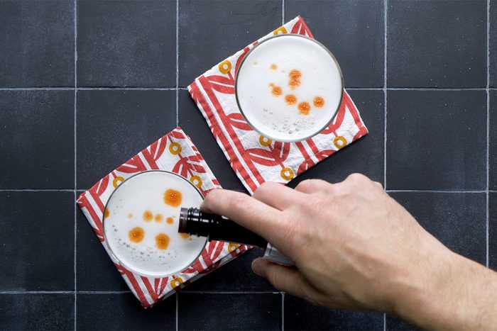 A hand adds drops of liquid, likely bitters, to the frothy surface of two cocktails served in glasses on red and white napkins.