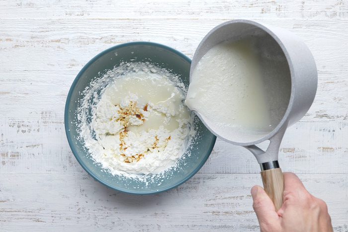 A person is pouring liquid from a saucepan into a mixing bowl containing flour and possibly other ingredients, on a white wooden surface.