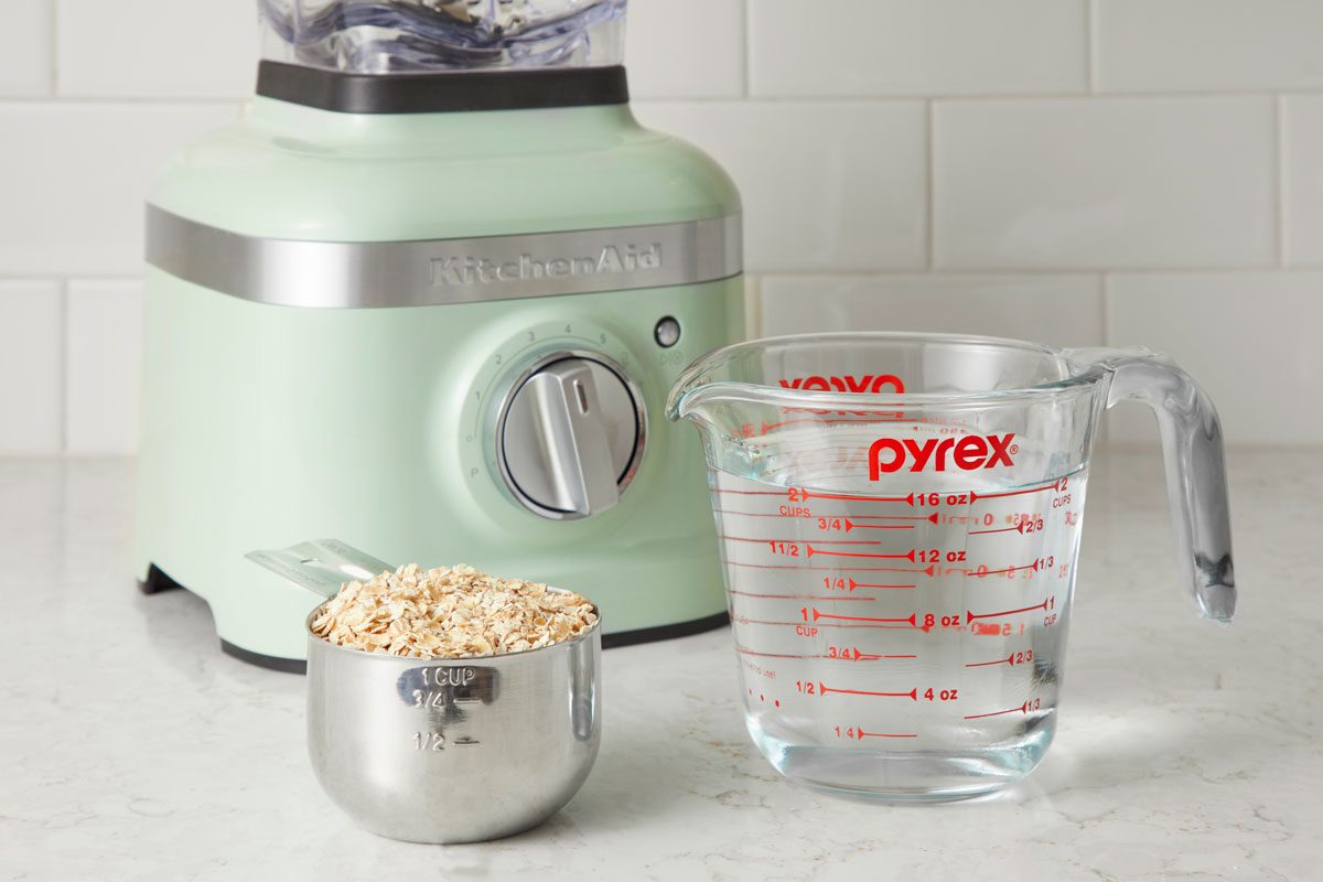 supplies for homemade oat milk on a kitchen counter; oats in a measuring cup near a measuring cup of water in front of a blender