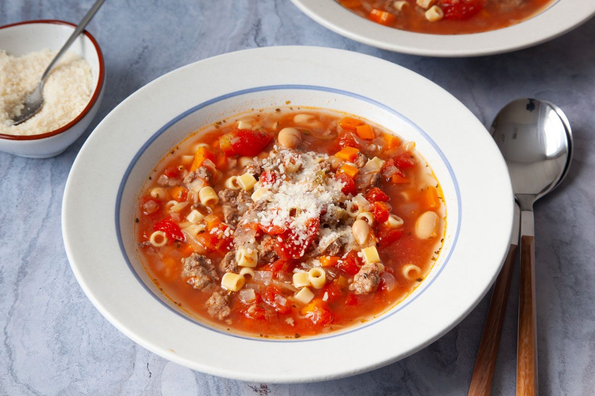 Single servings of Taste of Home Crockpot Pasta Fagioli is white bowls on blue gray surface with small bowl of parmesan cheese