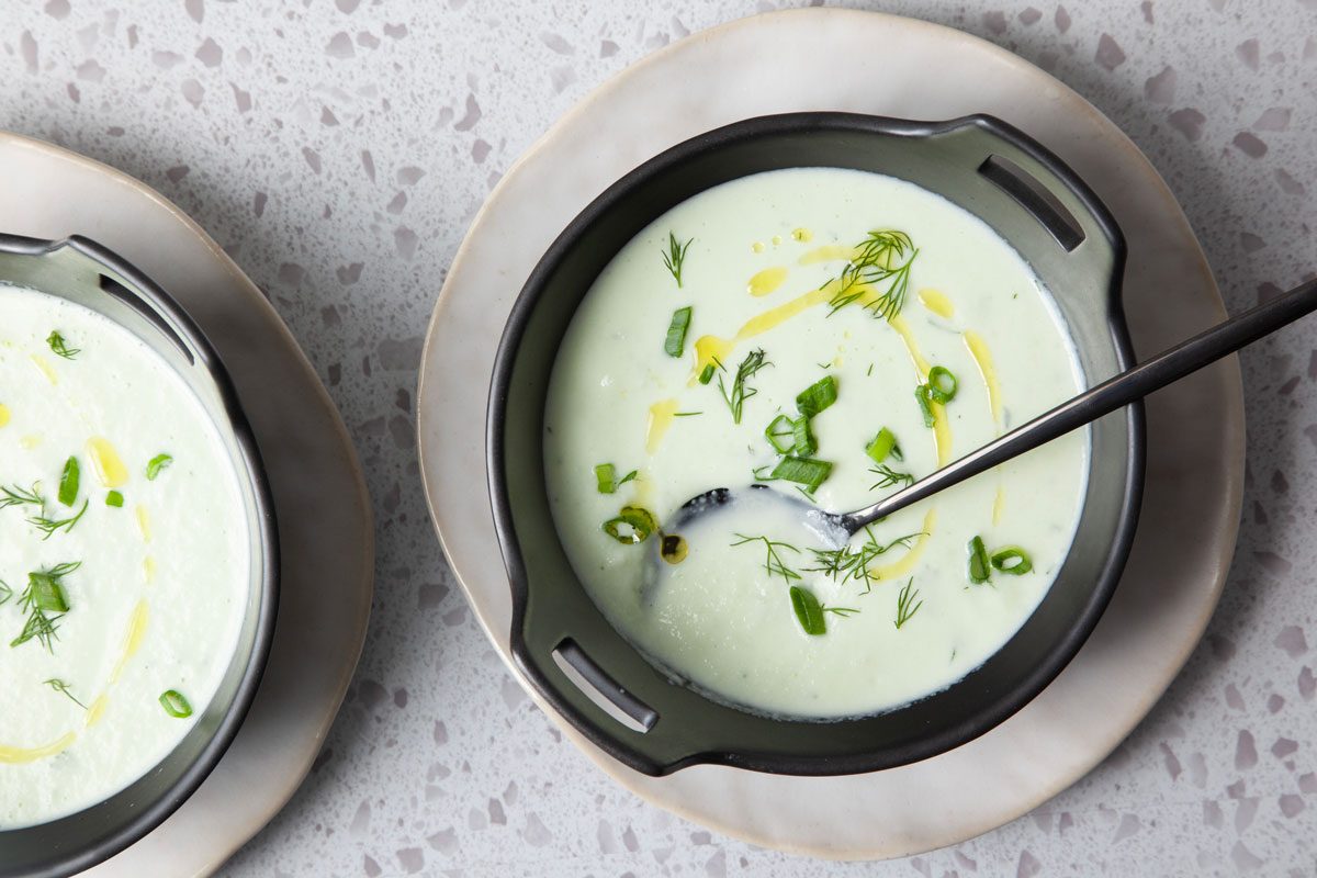 Single servings of Taste of Home Cucumber Soup in black bowls on a light gray plates, topped with olive oil, green onions and dill