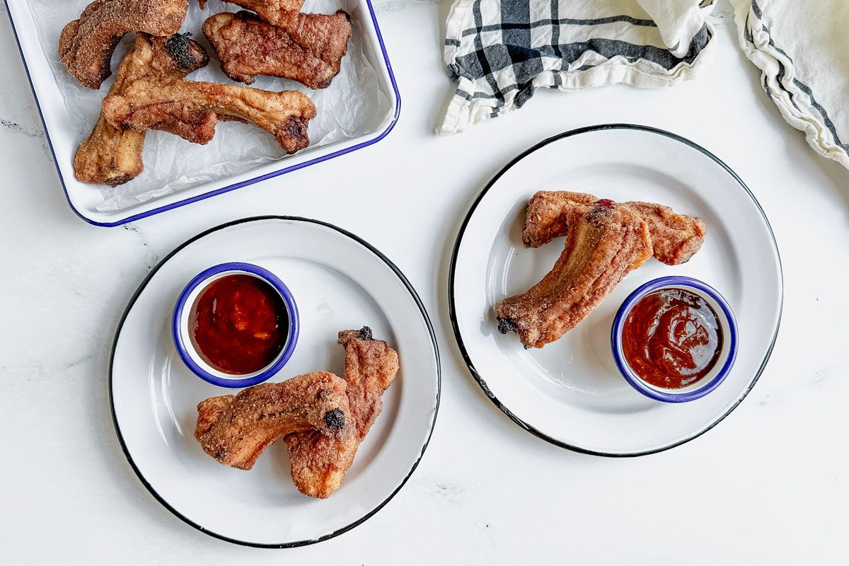 Taste of Home fried ribs on enamel trays and plates with barbecue sauce on a marbe surface next to a cotton napkin.