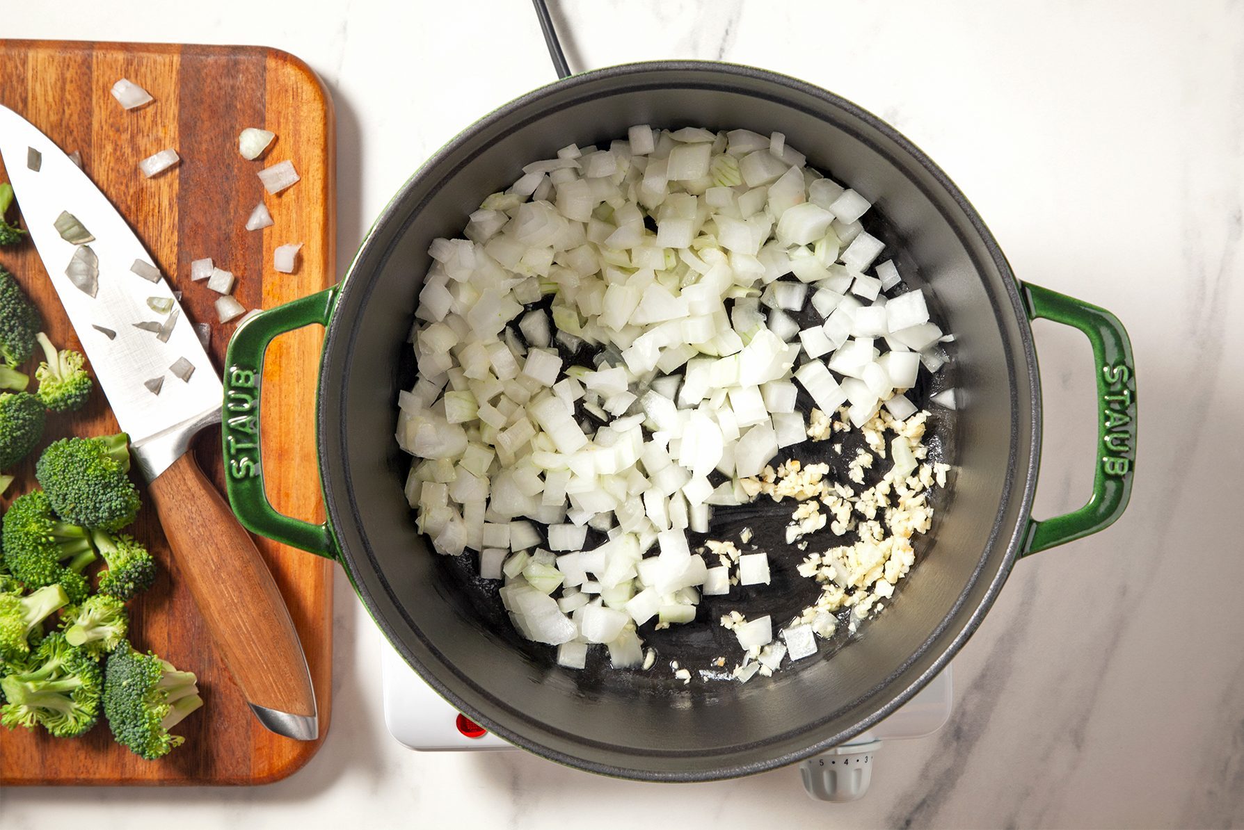 A pot on a stovetop with chopped onions and minced garlic sizzling inside. Next to the pot, there's a wooden cutting board with a knife, chopped onions, and broccoli florets. 