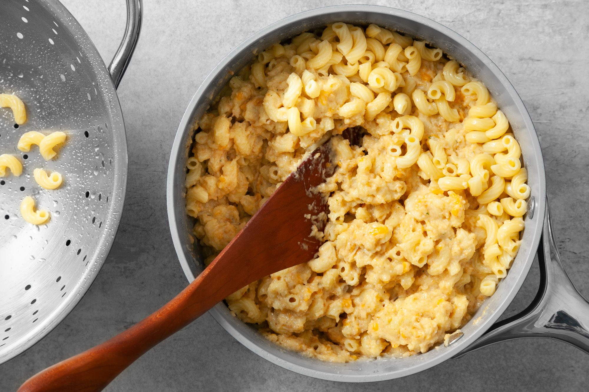 Overhead shot of same saucepan; add macaroni; toss to coat; wooden spatula; grey marble background;