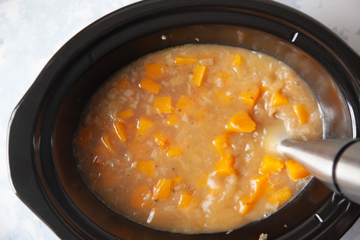 chopped butternut squash, potato, onion, garlic, fresh thyme, salt, pepper and chicken broth in a slow cooker, being pureed with an immersion blender