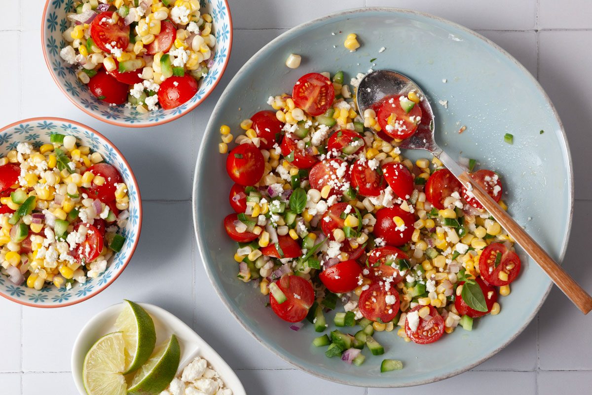 Full yield of Taste of Home Summer Corn Salad in a light blue bowl with feta and limes, 2 single servings in a blue and white bowls