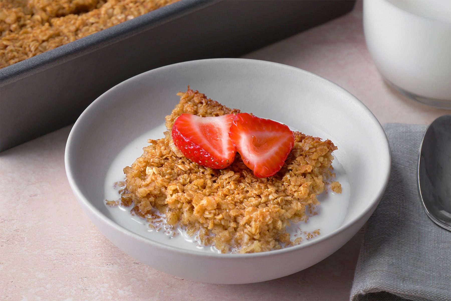 3/4th shot; light peach background; Baked Oatmeal served with milk on a white plate with silver spoon placed over grey kitchen towel, with a jar of milk;