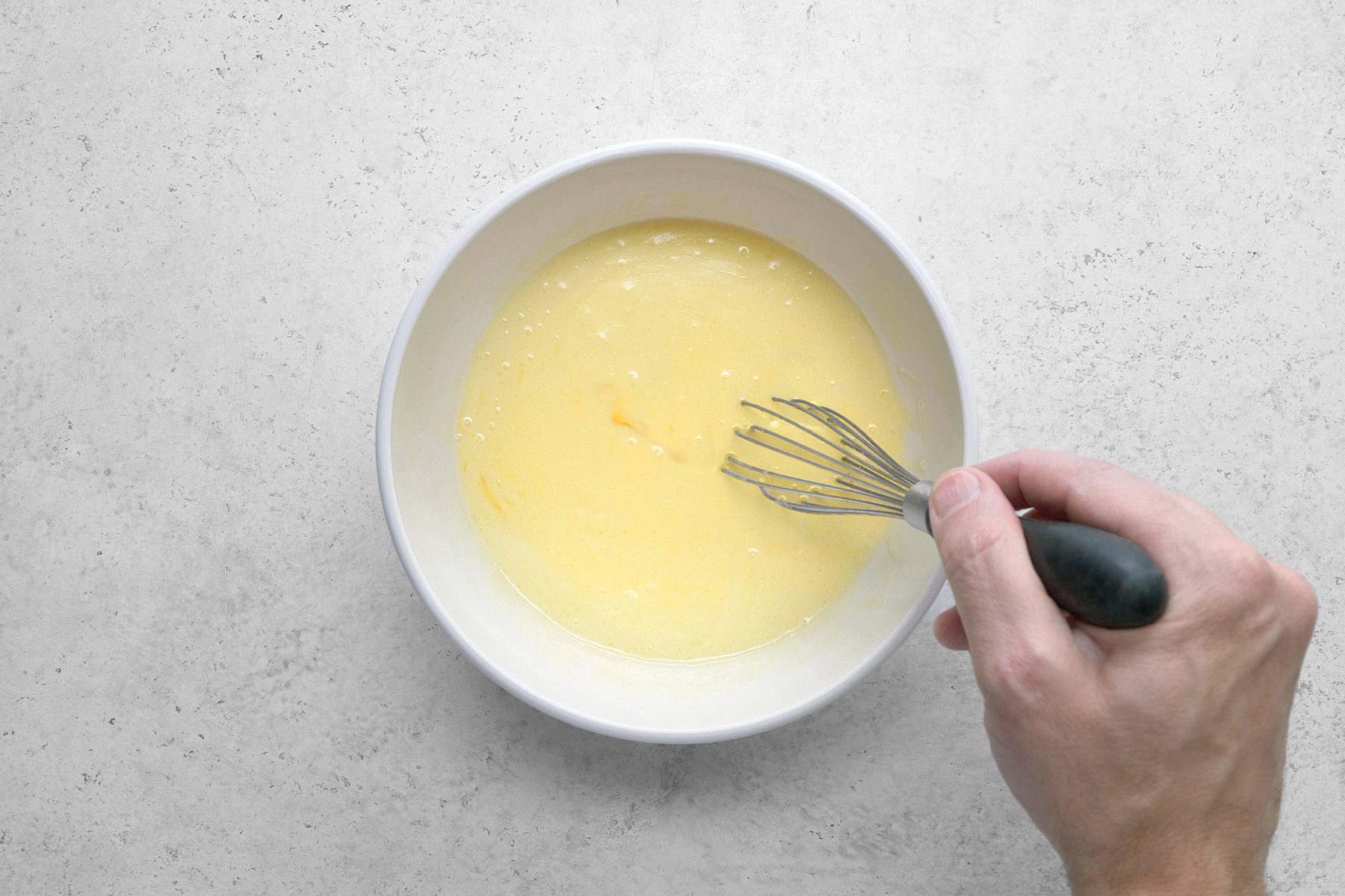 A person's hand whisking a yellow batter in a white bowl placed on a light gray countertop. The whisker has a black handle and the batter appears smooth. The scene suggests preparation for baking or cooking.