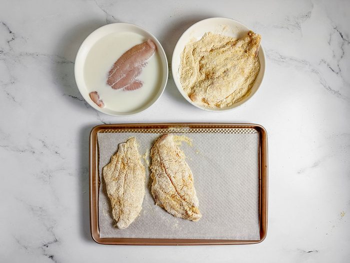 Breaded catfish fillets for Taste of Home catfish po-boy on a baking sheet next to breading ingredients in white bowls on a marble surface.