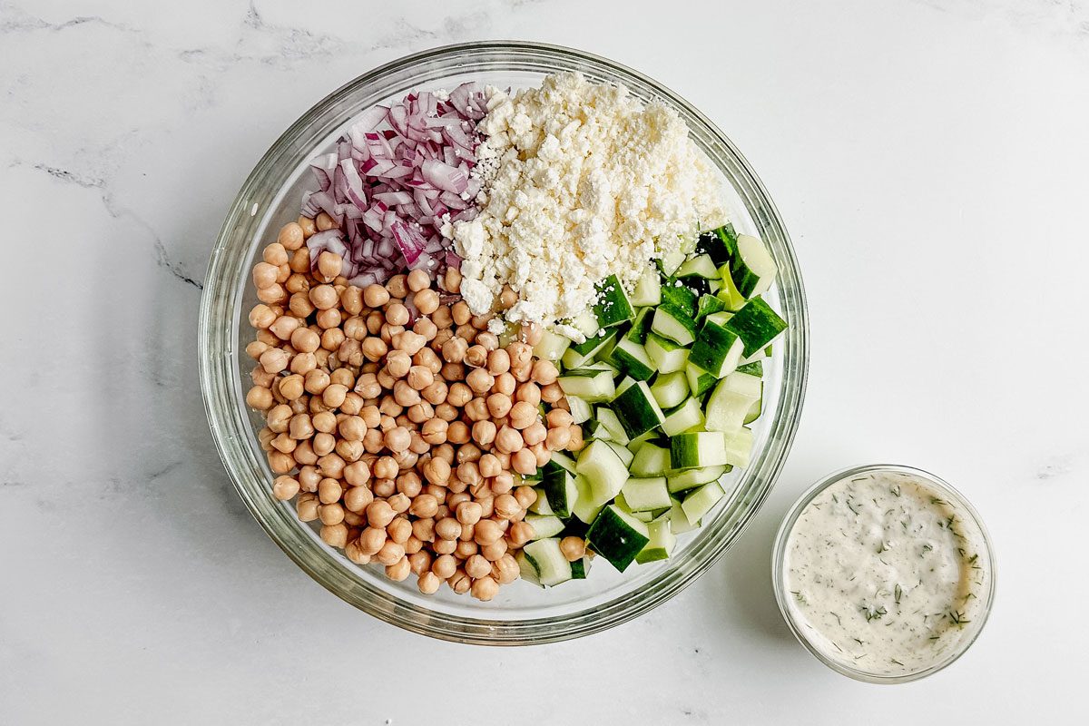 Ingredients for Taste of Home chickpea cucumber salad in a glass bowl on a marble surface.