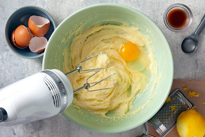 A mixing bowl filled with creamy batter and a cracked egg, being blended with an electric mixer. Nearby are broken eggshells in a small bowl, a zester with lemon zest, a spoon with vanilla extract, and a parchment paper on a gray countertop.