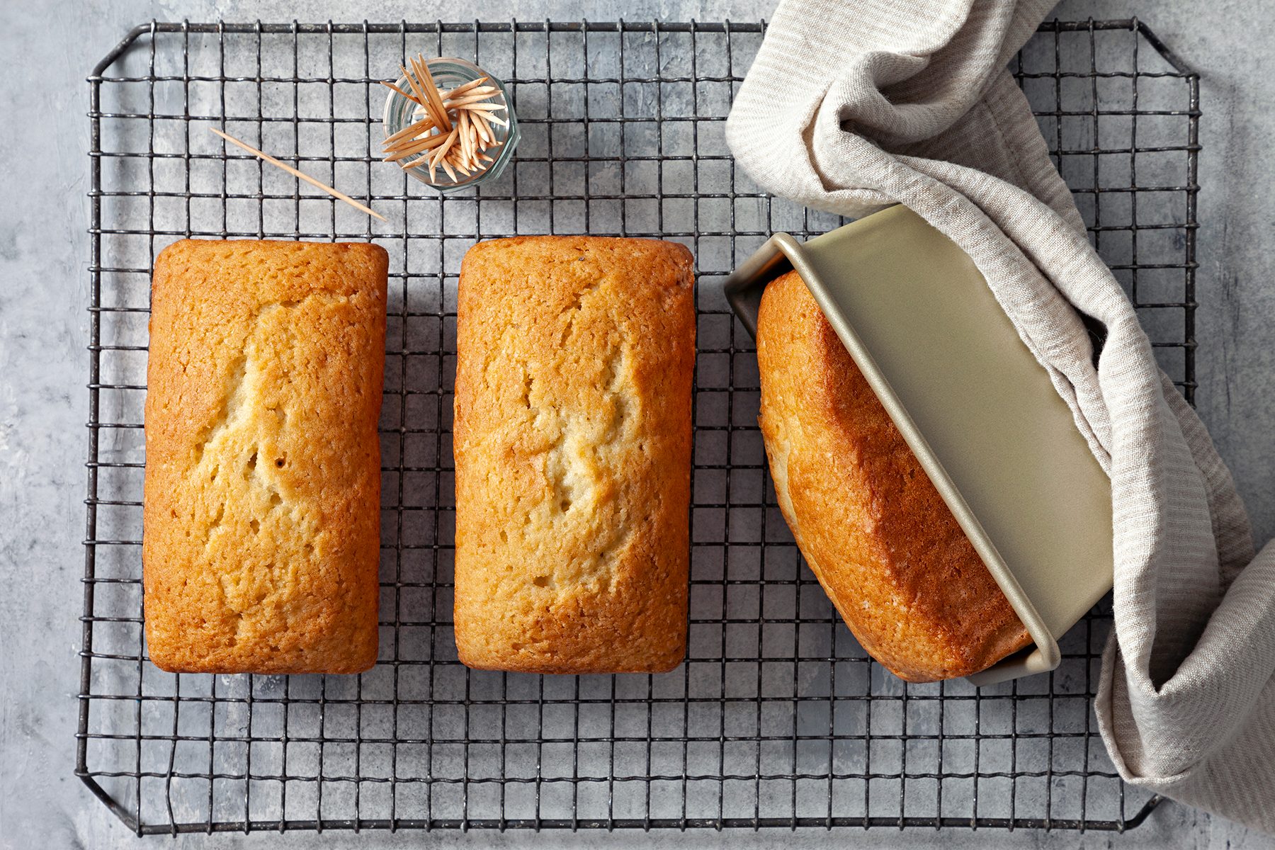 Three golden-brown loaves of bread rest on a cooling rack. Two are fully visible while the third is partially out of its baking pan, revealing a soft texture. 