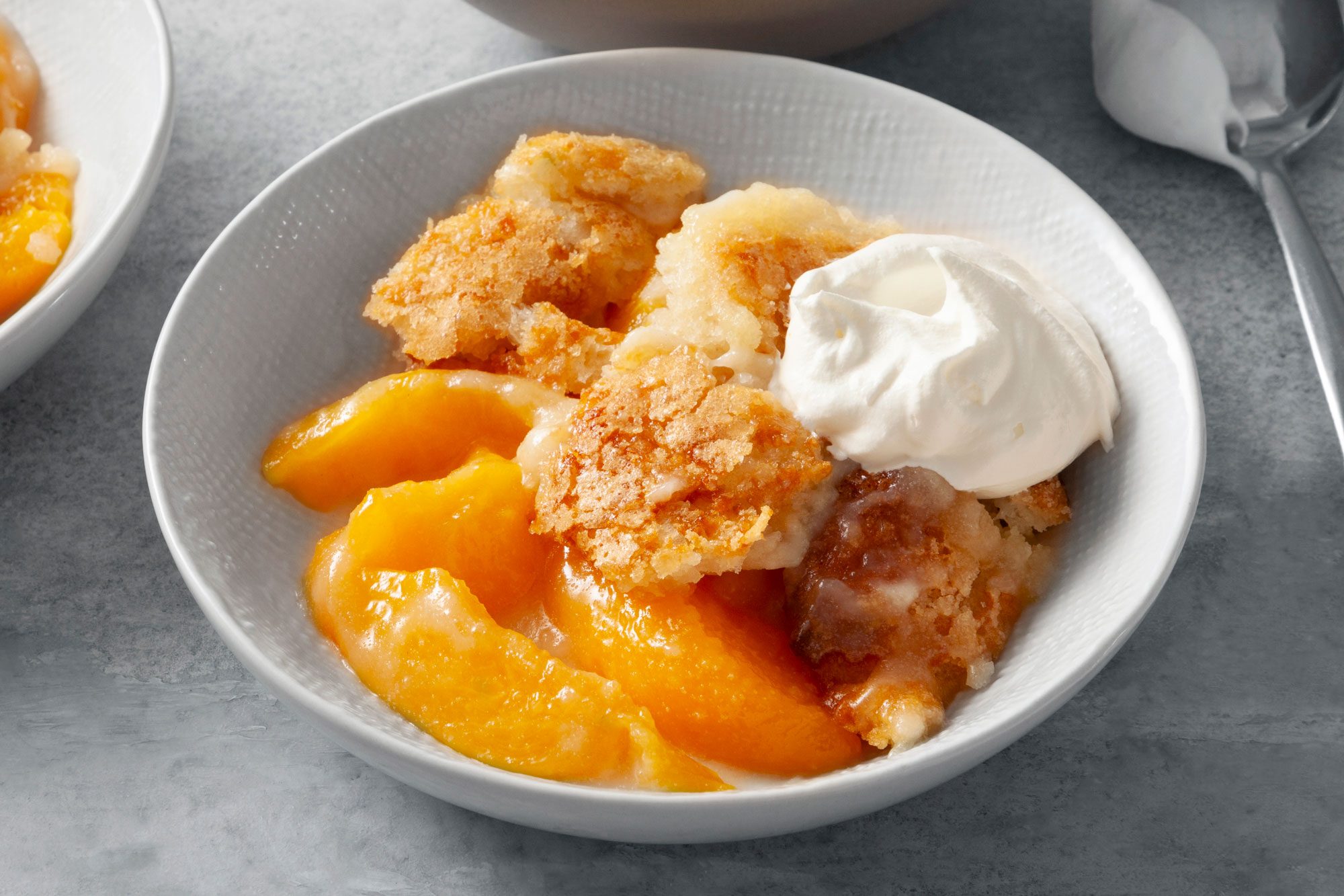Overhead shot of Peach Cobbler; served in bowl with whipped cream; spoon; grey marble surefcae;