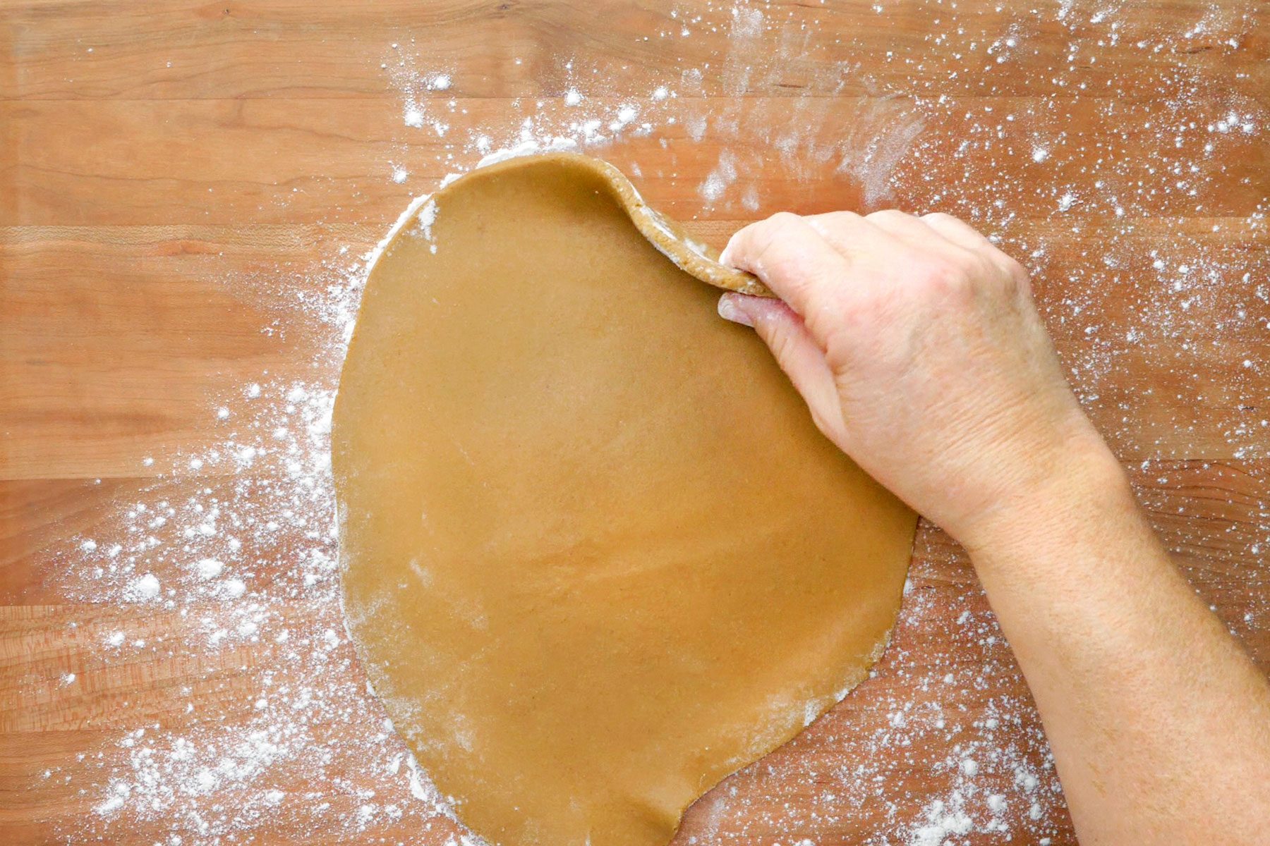 A hand lifting dough in the shape of a disk from a wooden surface.