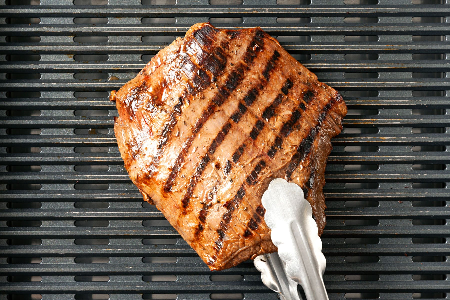 A large, perfectly grilled steak with visible char marks sits on a barbecue grill. A pair of tongs is positioned at one end holding the pice of steak.
