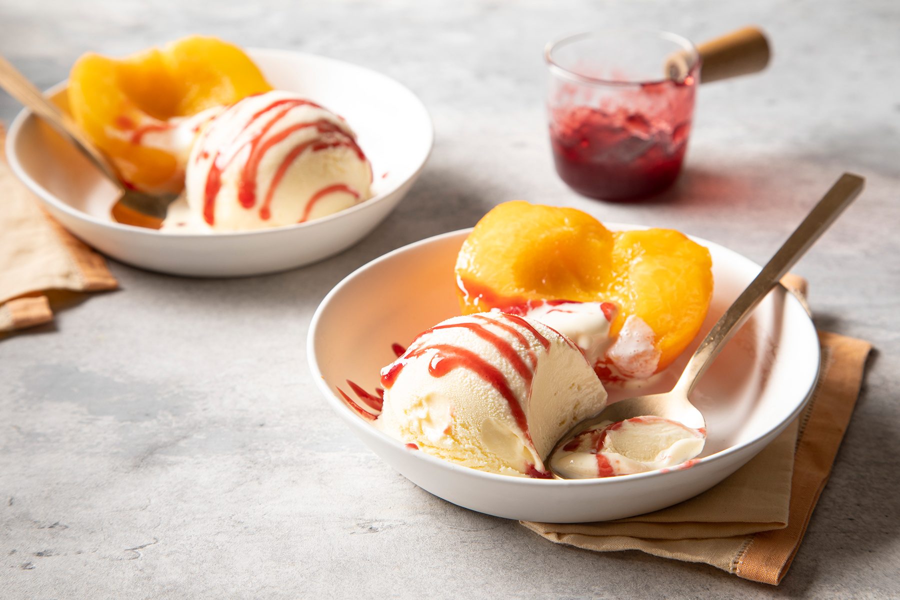 Two white bowls containing scoops of vanilla ice cream with peach halves and a drizzle of red sauce on top are set on a light gray surface. Each bowl has a spoon. A small glass container with red sauce and a wooden handle is visible in the background.
