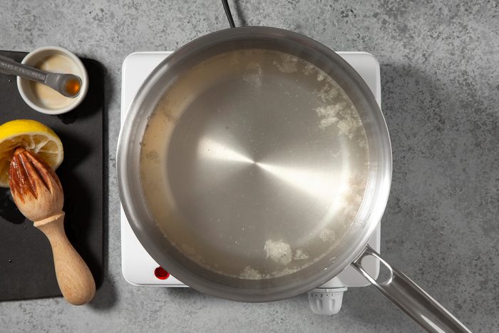 A top view of a silver saucepan with a metal handle on an electric stovetop. The pot contains a light, slightly translucent liquid. To the left, there is a cut lemon, a wooden citrus juicer, a small bowl with a brown liquid, and a spoon resting on a black surface.