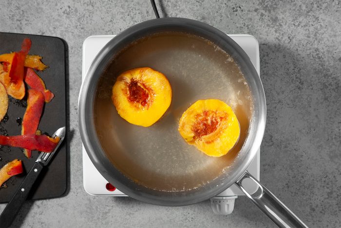 A small white stovetop with a pot of simmering water containing two halved peaches. To the left, a black cutting board holds a knife and peach peels. The surface is a gray, textured countertop.