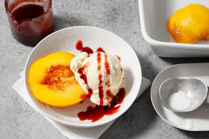 A dessert featuring a scoop of vanilla ice cream drizzled with red sauce, served beside a halved peach in a white bowl, on a light-colored surface with a jar of red sauce and another bowl containing a second halved peach and a measuring spoon.