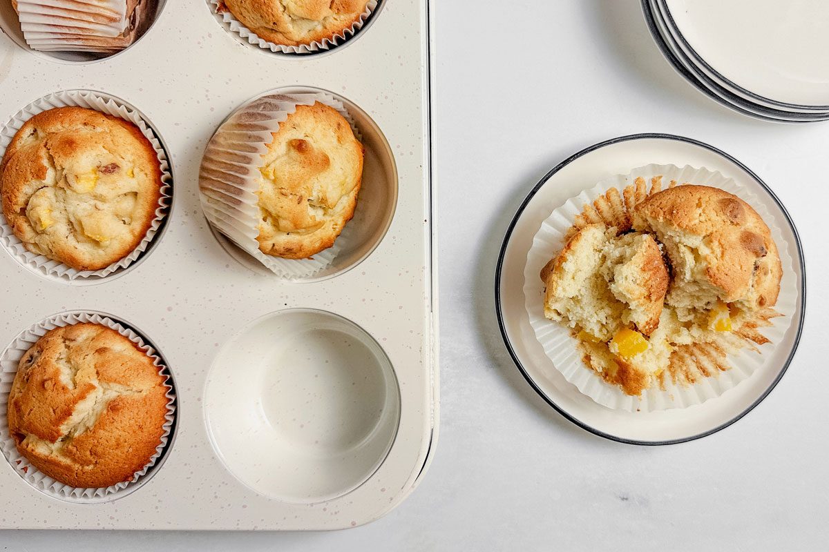 Taste of Home peach muffins in muffin tins and a white plate on a marble surface.