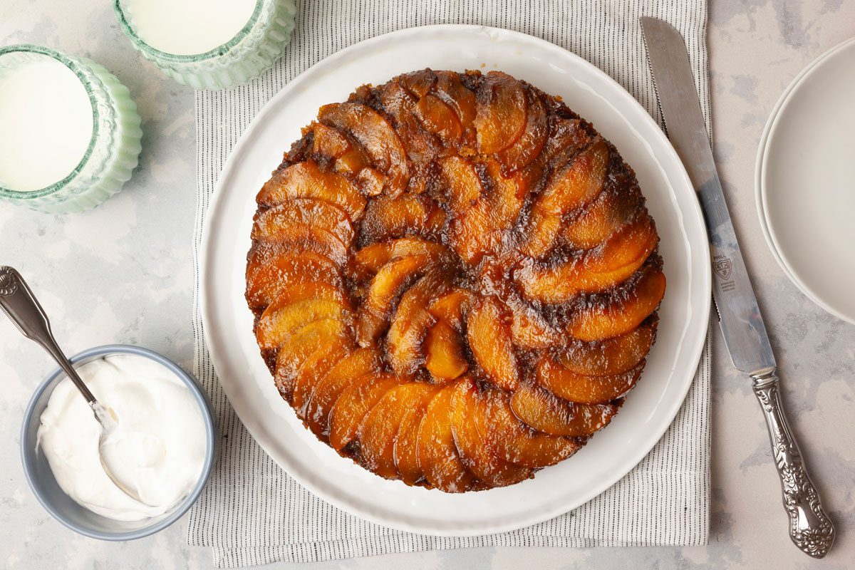Full yield of Taste of Home Peach Upside-Down Cake on a white platter, blue bowl of whipped cream