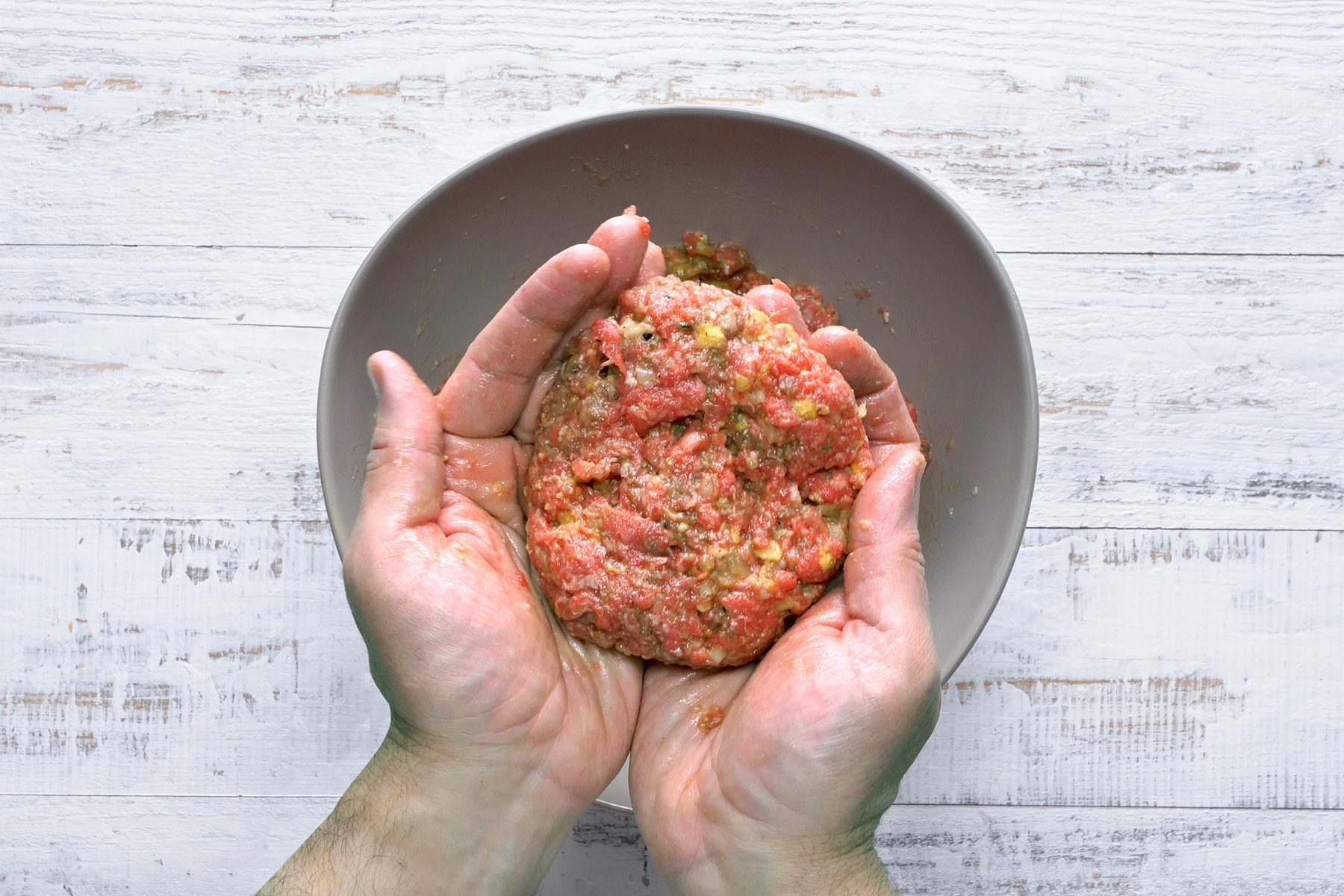 Hands shaping ground meat mixed with spices into a patty over a gray bowl with a rustic white wooden table as the background.
