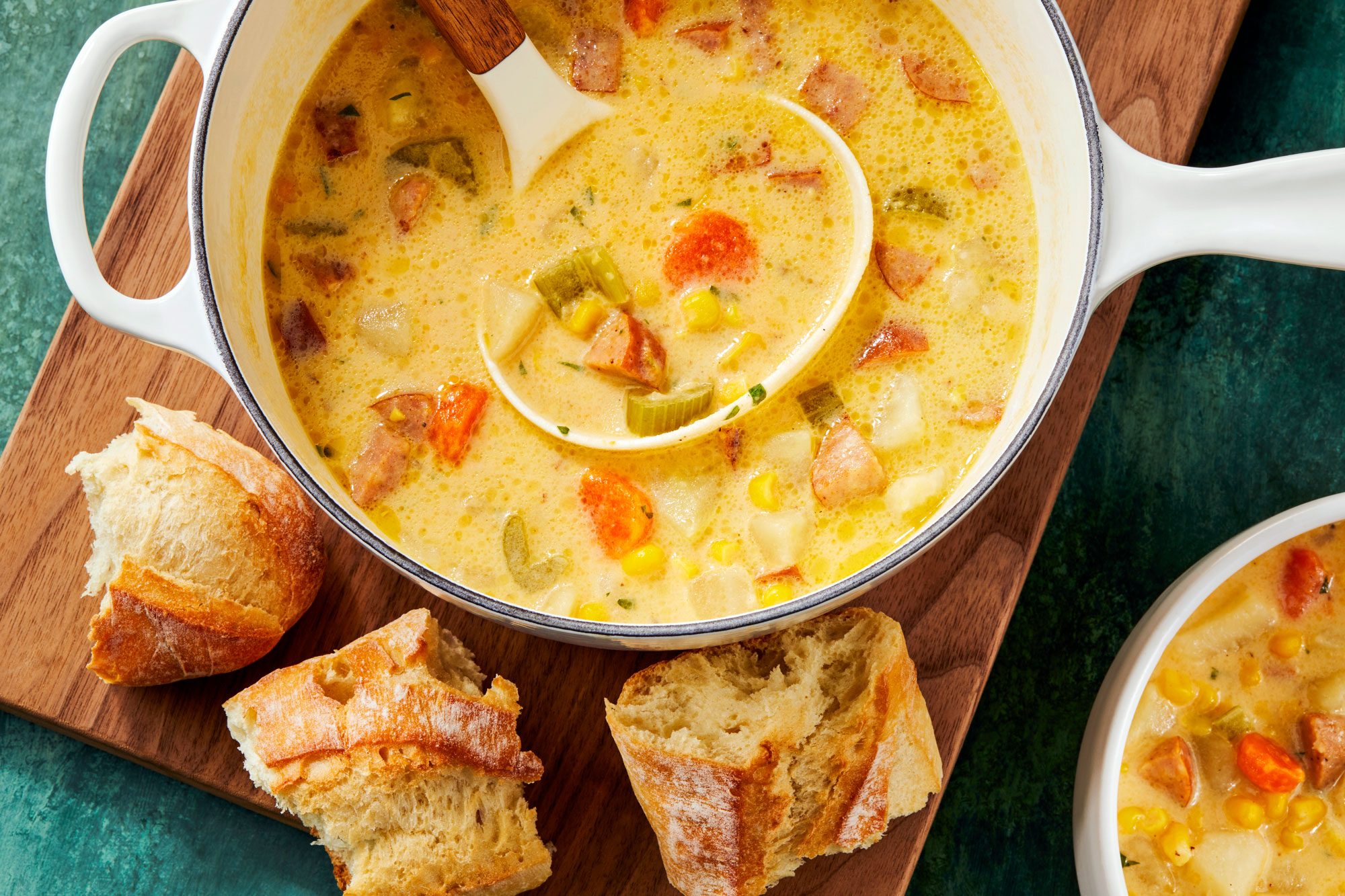 Overhead shot of Sausage Potato Soup; in a large saucepan on wooden board with breads; serve in a soup bowl; serving spoon; green texture surface;
