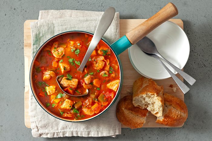 overhead shot; grey background; Seafood Soup in a pan over white kitchen towel with long serving spoon; two white plates with silver spoon and bread on wooden board