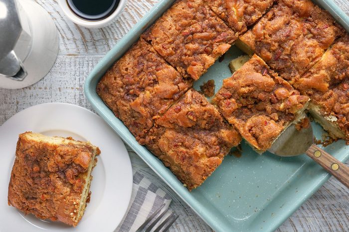 A top view of a baking dish filled with pieces of cake with a serving spatula. One piece has been placed on a white plate next to a cup of coffee, a fork, and a napkin, all on a light wooden surface.