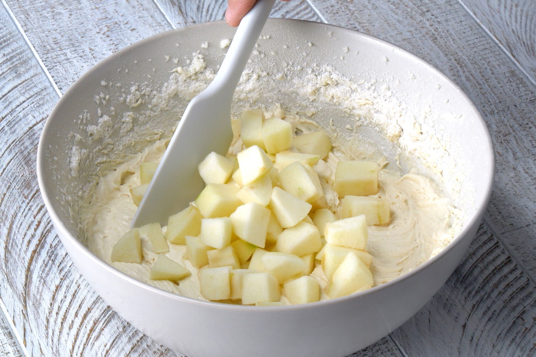 A rubber spatula is being used to add apples to the batter in a bowl.
