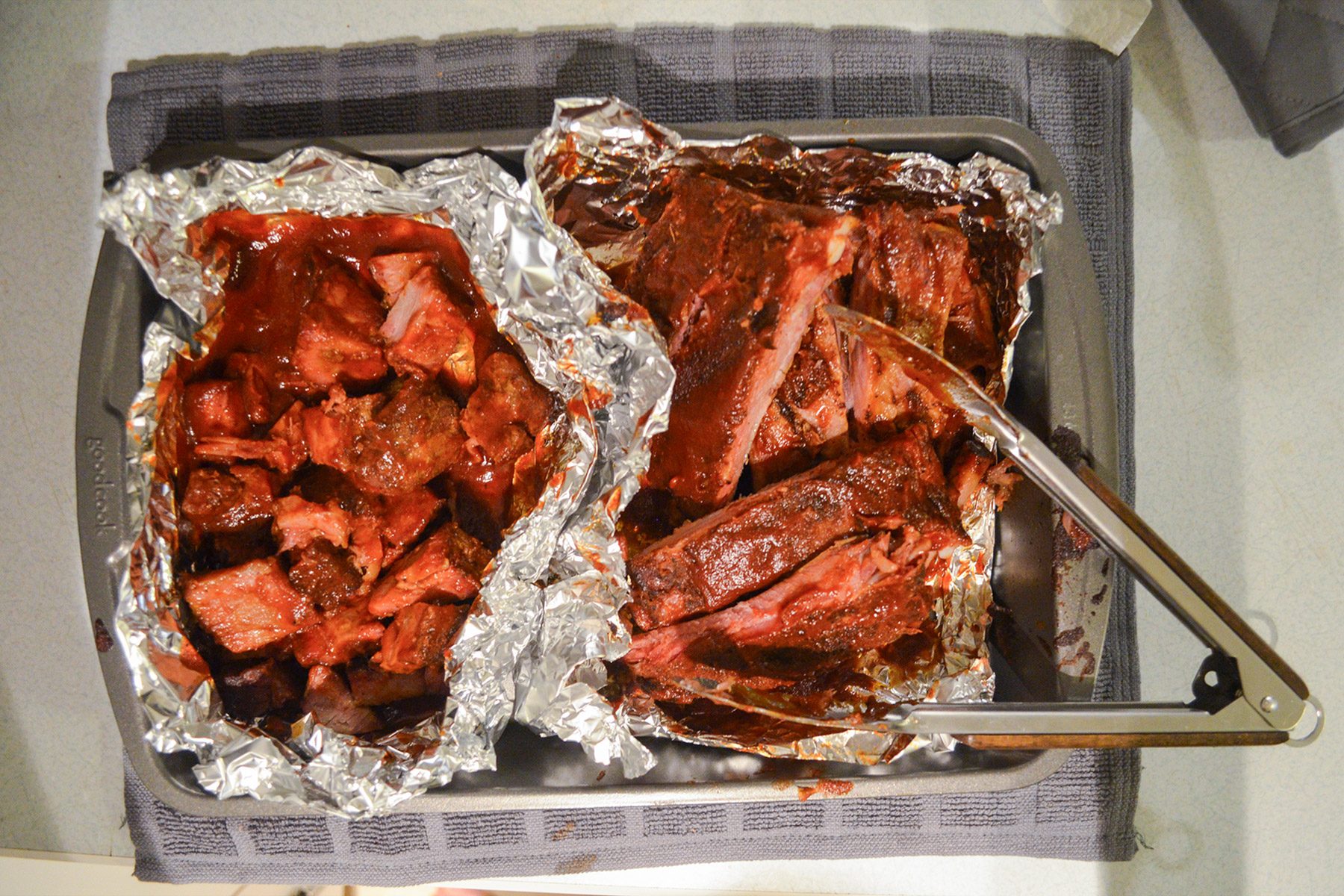 A tray contains two portions of foil-wrapped, barbecued meat. One side has cubed meat, while the other side has larger cuts. Tongs rest on the tray, and a gray kitchen towel is placed underneath it. The meat appears juicy and flavorful.
