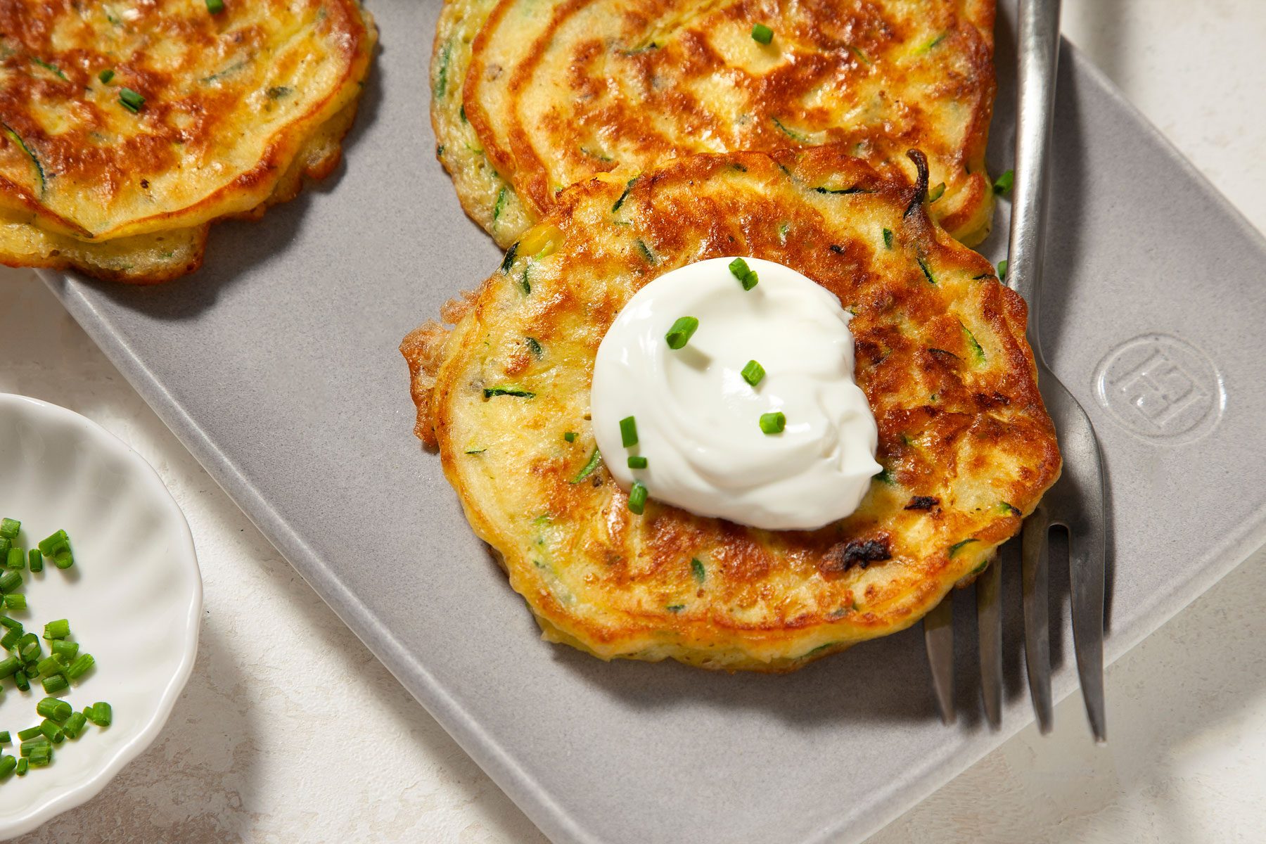 Two golden brown zucchini fritters topped with a dollop of sour cream and garnished with chopped chives on a silver tray. A fork rests beside the fritters.