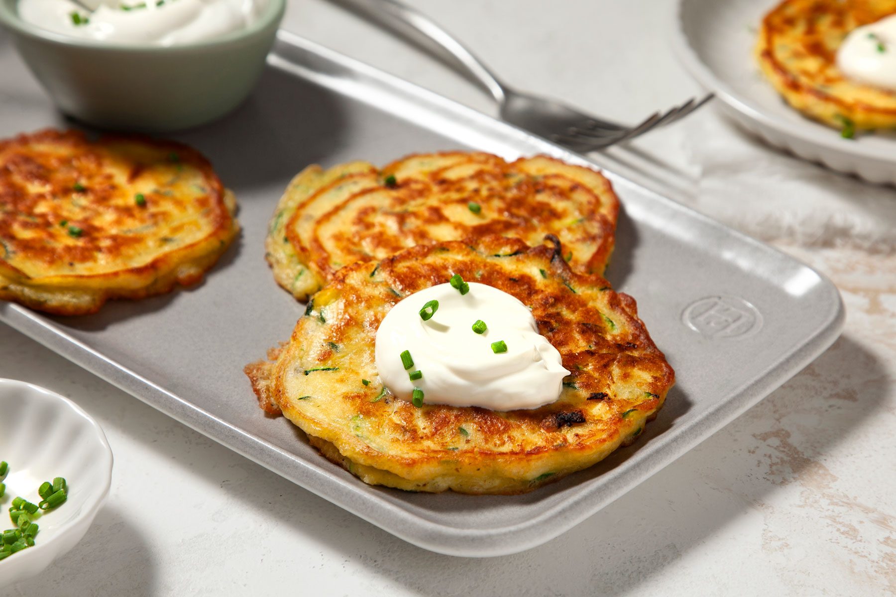 Two golden brown zucchini fritters topped with a dollop of sour cream and garnished with chopped chives on a silver tray. Nearby are additional dishes with fritters and a small white bowl containing more chives. A fork rests in the background.