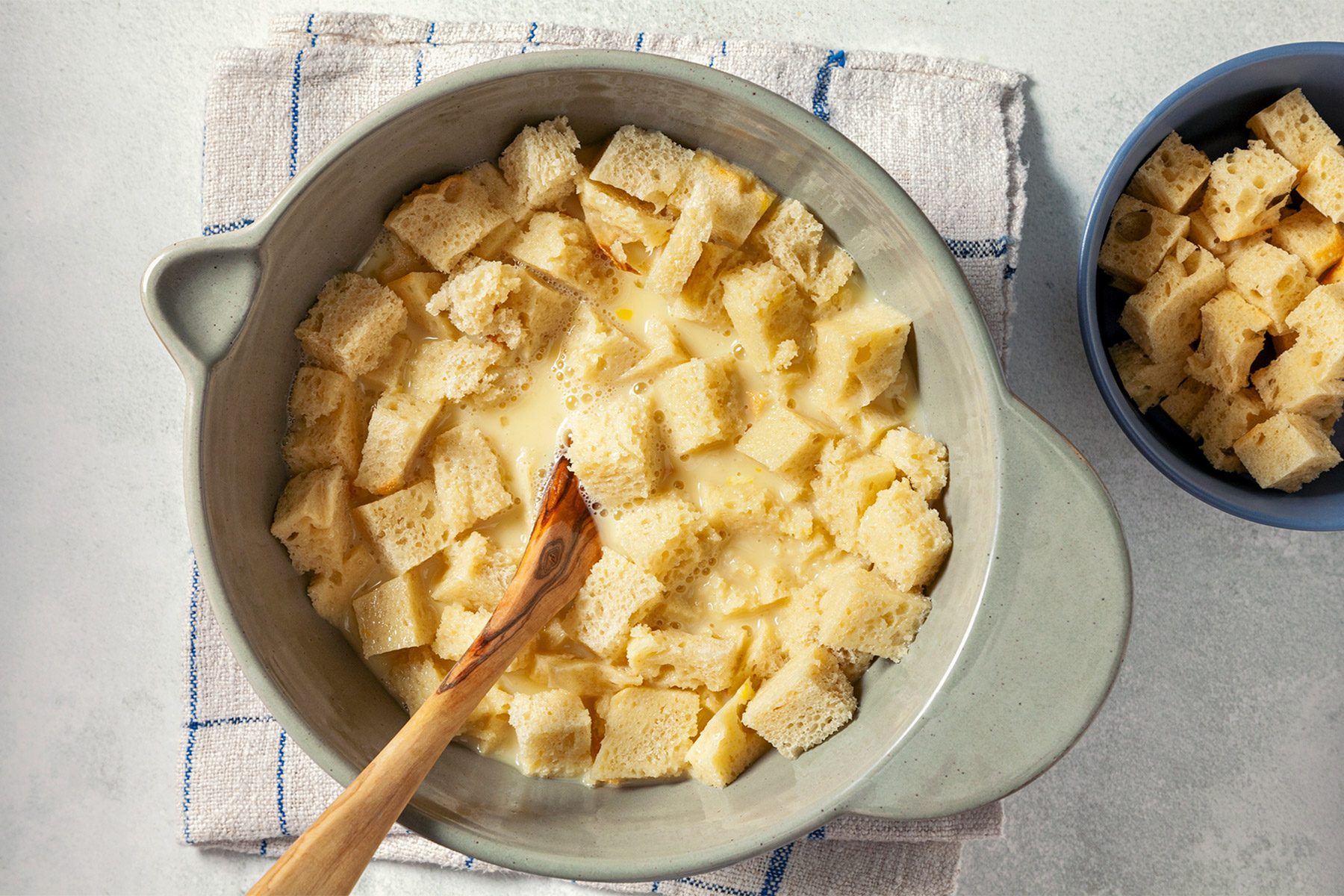overhead shot; white background; A grey bowl contains cubes of bread soaked in a creamy mixture; The bread cubes are partially submerged in the mixture; A wooden spoon rests in the bowl, A smaller bowl filled with additional bread cubes is in the background;