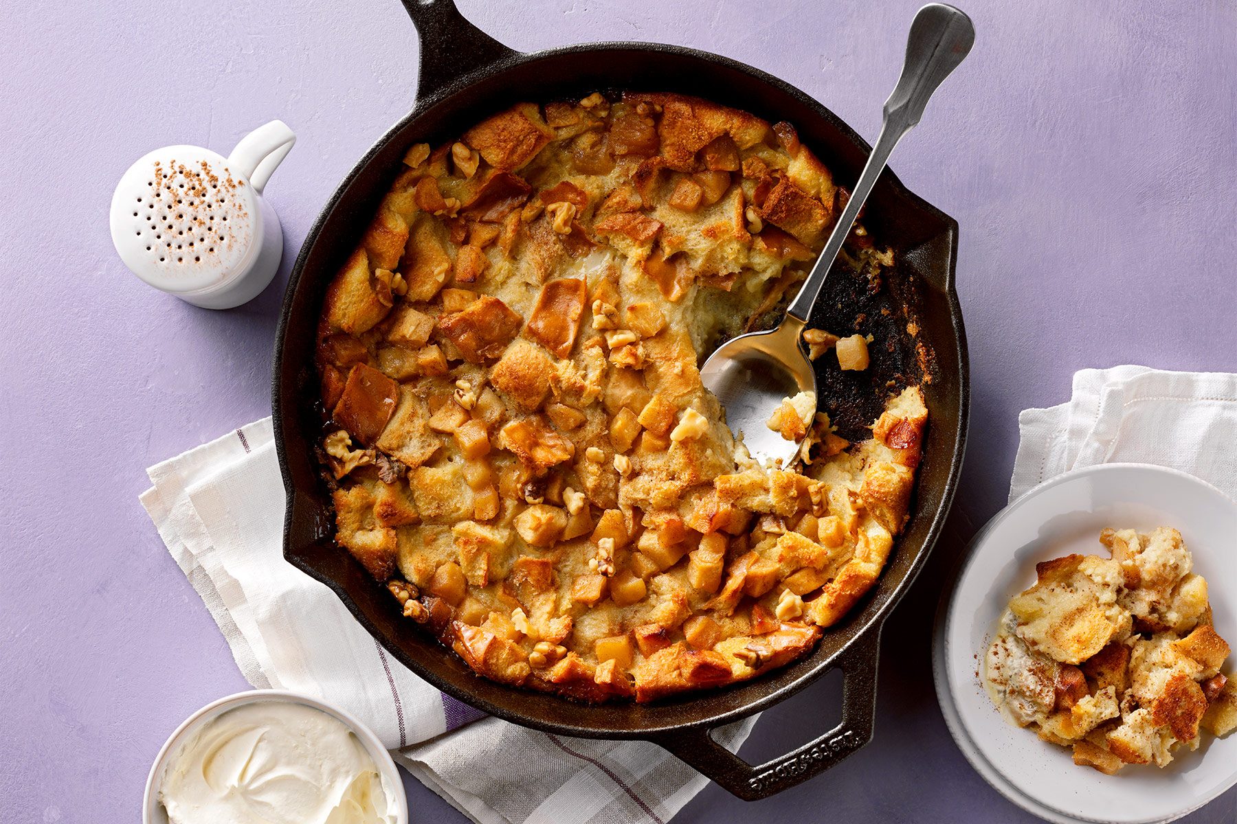 A cast-iron skillet filled with a golden-brown bread pudding. A spoon rests in the skillet. A white bowl containing a serving of the bread pudding is next to the skillet, along with a small container of cream on the left. The items are on a white cloth.