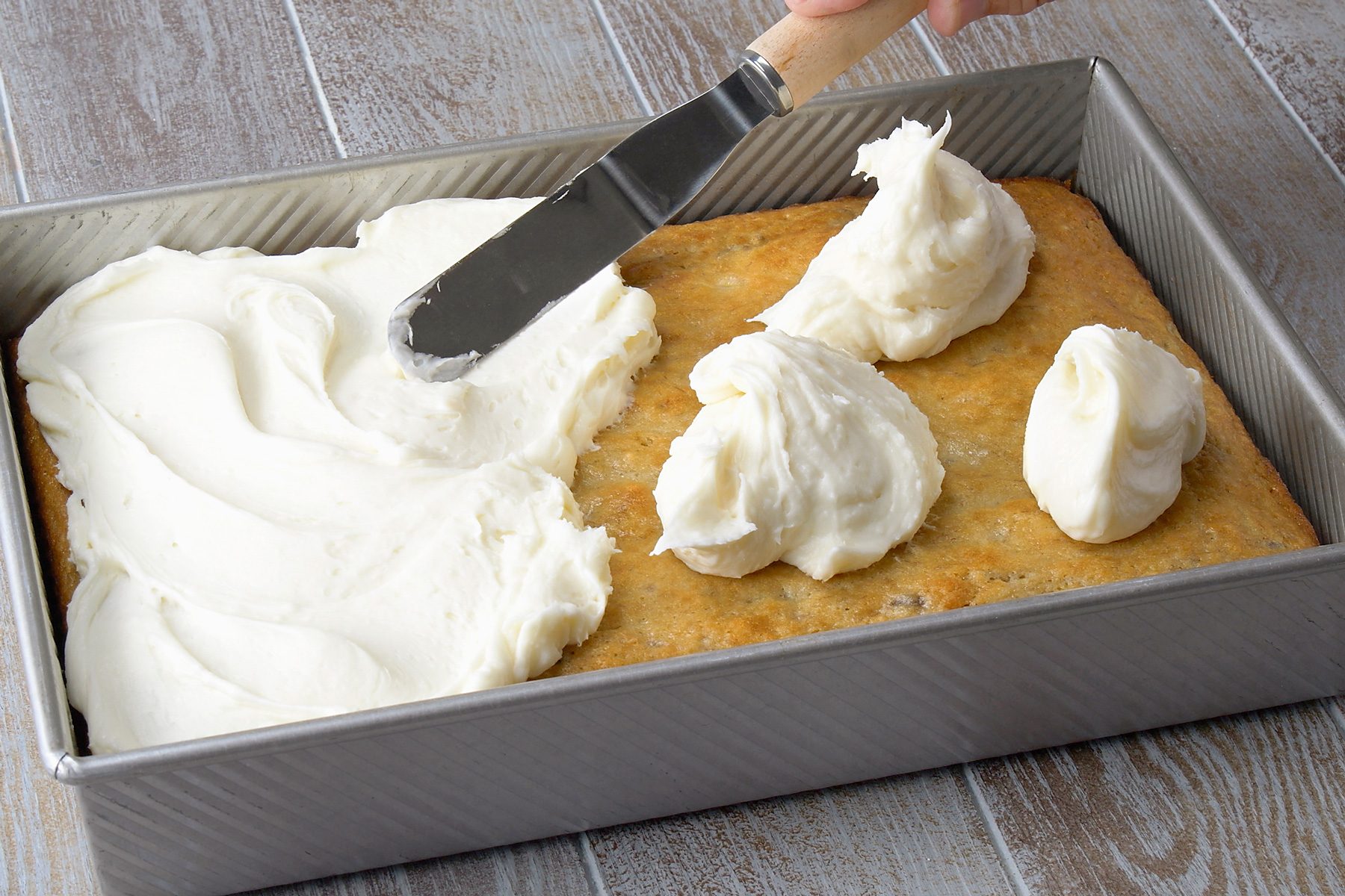 3/4th shot; grey background; A person is using an offset spatula to spread buttercream frosting onto a baked cake; The cake is in a rectangular baking pan, and the frosting is a creamy white color, The frosting is being spread evenly across the top of the cake; There are a few mounds of frosting on the cake, and some of the frosting is spilling over the edges of the pan