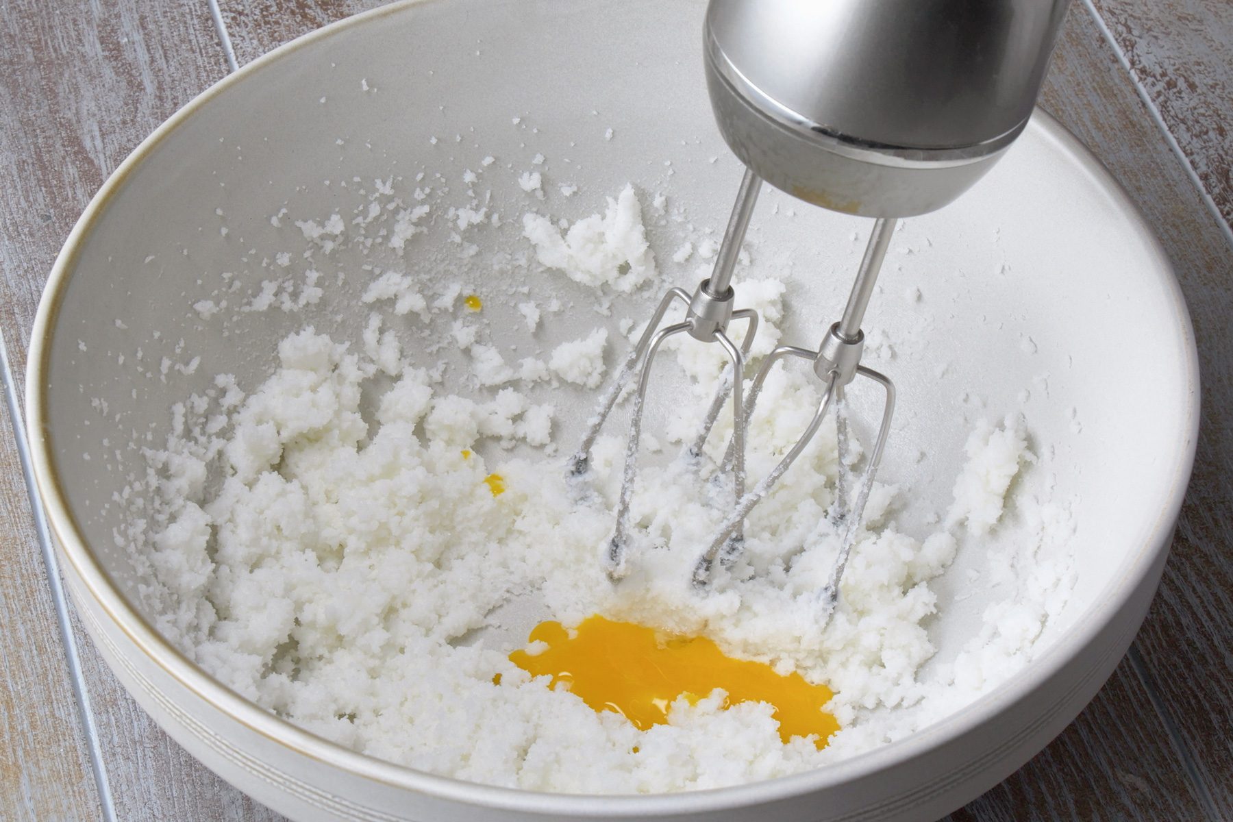 3/4th shot; grey background; A white bowl contains a mixture of cream cheese and sugar, An electric mixer is being used to beat the mixture together, The mixture is a pale yellow color and is starting to become light and fluffy, A single egg yolk is visible in the bowl